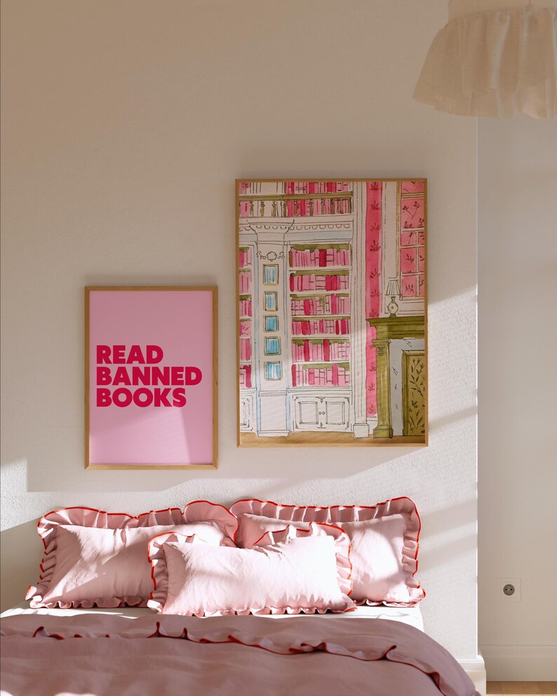 May include: A bedroom scene featuring a bed with pink bedding and pillows with red trim. Two framed artworks hang on the wall: one with the text "READ BANNED BOOKS" and another depicting a library interior. The room is painted in soft pink tones.
