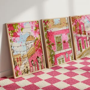 May include: Three framed watercolor prints depict a European street scene with pink buildings, green accents, and a domed structure. The artwork is in a light wood frame, and the scene is set against a pink and white checkered rug.