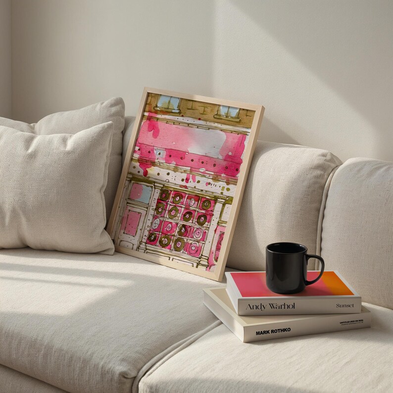 May include: A framed watercolor print with a pink and gold abstract design. The print is propped on a white couch next to a black mug and two books. The books are titled "Andy Warhol" and "Mark Rothko".