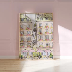 May include: Watercolor print of a Parisian street scene with the Eiffel Tower, buildings with flower boxes, and outdoor cafes with pink umbrellas. The artwork is framed in a gold-colored frame and leans against a pink wall.