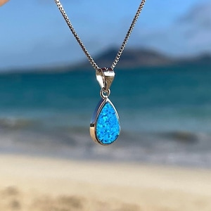 May include: A silver chain necklace with a teardrop-shaped pendant featuring a blue opal gemstone. The necklace is photographed against a beach background.