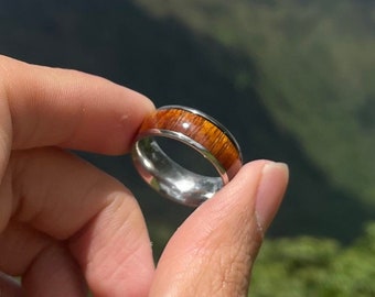 May include: A silver ring with a brown wood inlay. The ring is being held up in front of a mountain range and a blue sky with white clouds.