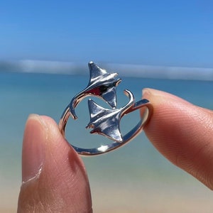 Puede incluir: Un anillo de plata con dos diseños de mantarrayas entrelazados. El anillo se muestra en un dedo contra un fondo de playa.