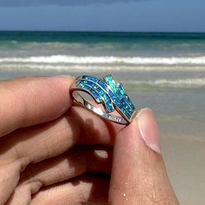 Könnte beinhalten: Ein silberner Ring mit wellenförmigem Design, besetzt mit blauen und türkisfarbenen Steinen. Der Ring wird vor dem Hintergrund eines Sandstrandes und Ozeans unter blauem Himmel gehalten.