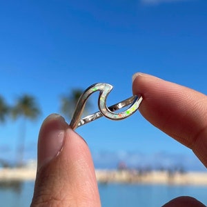 May include: A silver wave ring with an iridescent opal inlay. The ring is being held by a hand against a blue sky and ocean background.