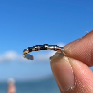 May include: A silver-colored toe ring with a bamboo design. The ring is held up against a bright blue sky and ocean background. The ring is small and delicate, with a textured surface.