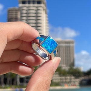 May include: A silver ring with a square blue opal gemstone. The ring is being held by a hand in front of a blurry background of buildings and a body of water.