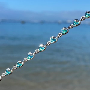 Pode incluir: Uma delicada pulseira de corrente prateada com um padrão repetitivo de pequenos berloques turquesa em forma de onda. A pulseira é apresentada em um fundo de céu azul brilhante e oceano, sugerindo um tema de praia ou costeiro.