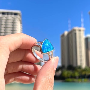 May include: A silver ring with a triangular blue opal gemstone. The ring is held in a hand with a blurred background of a beach and buildings.