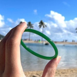 May include: A vibrant green jade bangle bracelet held up against a tropical beach scene. The smooth, circular bracelet is the focal point, with a clear, glossy finish. The background features a blue sky, palm trees, and a sandy beach.
