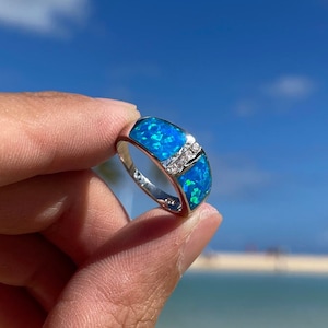 Puede incluir: Un anillo de plata con una piedra de &oacute;palo azul y acentos de circonita c&uacute;bica transparentes. El anillo se coloca sobre un fondo de cielo azul y oc&eacute;ano.