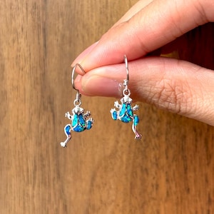 May include: A pair of silver-toned frog earrings with turquoise blue accents. The earrings are held against a wooden background. The frog design features detailed legs and a textured body, creating a whimsical accessory.