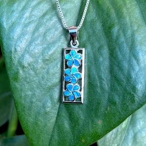 May include: A silver necklace with a rectangular pendant featuring three blue flower designs. The pendant hangs from a delicate chain, set against a backdrop of large green leaves. The flowers have a detailed, layered appearance.