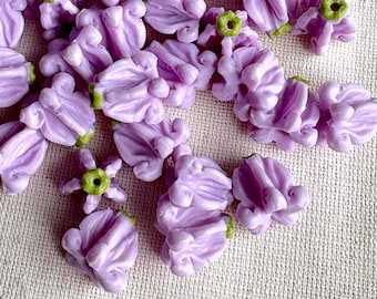 May include: A collection of small, light purple flower-shaped beads with green accents. The beads are clustered together, showcasing their detailed floral design. These beads are likely for jewelry making or craft projects.