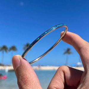May include: A silver bangle bracelet with a blue and green opal inlay. The bracelet is being held up against a blue sky with palm trees in the background.