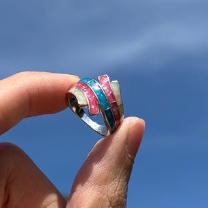 May include: A silver ring with pink and blue enamel detailing. The ring is on a finger against a blue sky background.