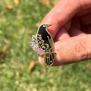 May include: A black enamel pin featuring a crow perched on a branch. The crow has gold accents and is decorated with a crescent moon, stars, and floral details in shades of purple, pink, and blue. The pin is held against a background of green grass.