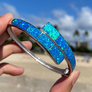 Könnte beinhalten: Ein silbernes Armreif-Armband mit einem blauen Opal-Inlay. Das Armband besteht aus kleinen, quadratischen Opalen, die in einem silbernen Band eingefasst sind. Das Armband wird auf einer Hand vor einem Strandhintergrund gezeigt.