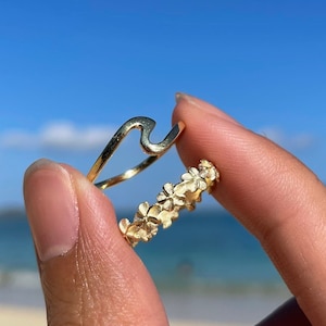 May include: Two gold rings, one shaped like a wave and the other with a floral design, are worn on a finger.
