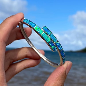 May include: A silver bangle bracelet with a blue and green opal inlay. The bracelet is open and has a simple design.