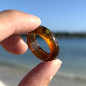 May include: A close-up of a brown and green resin ring with a unique marbled pattern. The ring is being held by a hand against a beach background.