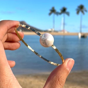 May include: A gold bangle bracelet with a large white pearl charm. The bracelet is shown against a beach background.