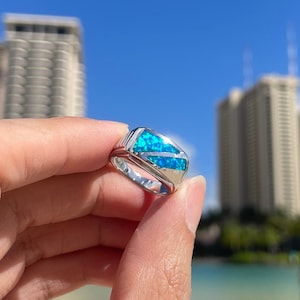 May include: A silver ring with a blue opal stone set in a square shape. The ring is being held by a hand in front of a blurry background of buildings and water.