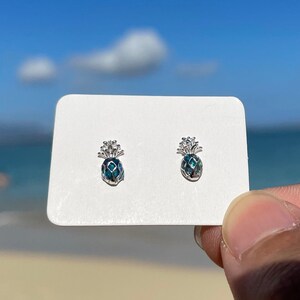 Op de afbeelding: Een paar zilveren ananas oorbellen met blauwe en groene abalone schelp inlegwerk. De oorbellen worden gepresenteerd op een witte kaart.
