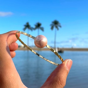 Może przedstawiać: Złota bransoletka z pojedynczą różową perłą w środku. Bransoletka jest trzymana w dłoni na tle plaży i palm.