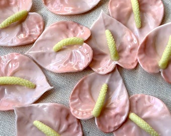 May include: A collection of pink ceramic anthurium flower shapes with yellow stamens. The flowers are arranged on a light-colored fabric surface. The anthuriums have a glossy finish and are in various stages of bloom.