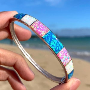 May include: A silver bangle bracelet with blue, pink, and white opal inlay. The bracelet is shown on a hand against a beach background.