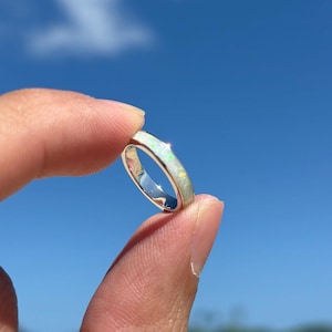 Puede incluir: Un anillo de plata con una banda de material blanco e iridiscente similar al ópalo. El anillo se sostiene entre dos dedos sobre un fondo de cielo azul brillante y océano.