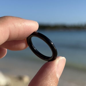 May include: A black ring made of a smooth, shiny material. The ring is held in a hand against a background of a beach and blue sky.