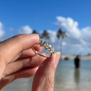 May include: A gold ring with a cluster of small, clear stones set in a band. The ring is held in a hand against a background of a beach and blue sky.
