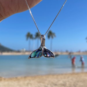 May include: A silver chain necklace with a blue and green abalone shell whale tail pendant. The necklace is being held by a hand in front of a beach scene.