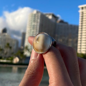 May include: A silver ring with a white oval-shaped gemstone. The ring is set against a backdrop of a beach and buildings.