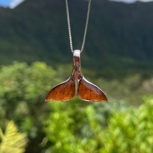 May include: A silver chain necklace with a brown whale tail pendant. The pendant is made of wood and has a natural grain pattern. The necklace is photographed against a blurred background of green foliage and mountains.