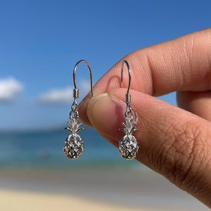 May include: A pair of silver pineapple earrings with a dangling design. The earrings are held in a hand against a blue sky and beach background.