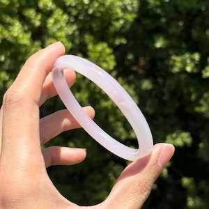 May include: A pale pink, translucent bangle bracelet. The smooth, rounded bracelet is held against a backdrop of green foliage. The light catches the bracelet, highlighting its delicate colour and form. This piece is a type of jewellery.