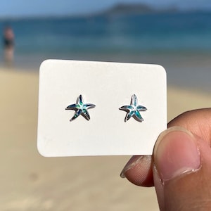 Può includere: Un paio di orecchini a forma di stella marina argentati con accenti iridescenti verdi e blu. Gli orecchini sono esposti su una carta bianca.