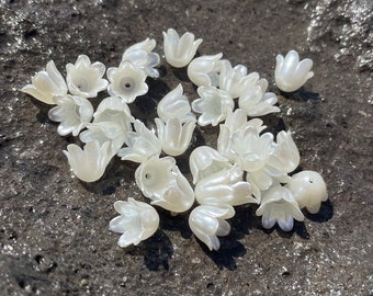 May include: A collection of white, pearl-like flower-shaped beads. The beads are scattered on a dark, textured surface.