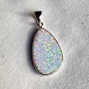 May include: A pear-shaped pendant with a shimmering, iridescent opal stone set in a silver-colored metal frame. The opal displays a range of colors, including pink, green, and yellow, against a white background. The pendant is displayed on a white fabric.