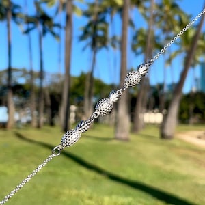 May include: A silver chain bracelet with four pineapple charms. The bracelet is held in a hand against a blurred background of palm trees.