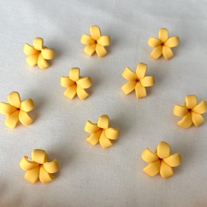 Op de afbeelding: Een verzameling kleine, gele bloemversieringen met vijf bloemblaadjes. De bloemen zijn verspreid over een wit oppervlak, met een aaneengesloten streng van vergelijkbare bloemen aan de onderkant. De bloemen hebben een zacht, afgerond uiterlijk.