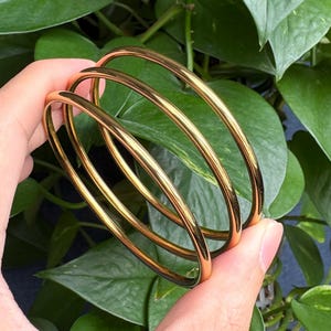 May include: Three gold-colored bangles are held in a hand, set against a backdrop of lush green leaves. The bangles are smooth and shiny, reflecting light. The jewelry is simple and elegant.