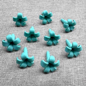 May include: Nine turquoise flower-shaped beads are arranged on a gray textured surface. Each bead has a central point with curved petals, resembling small blossoms. The beads are evenly spaced, creating a visually appealing pattern.