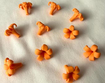 Könnte beinhalten: Eine Ansammlung kleiner, orangefarbener, blütenförmiger Verzierungen. Die Miniaturblüten sind auf einer weißen Oberfläche angeordnet. Die Blumen haben ein detailliertes, realistisches Design mit Blütenblättern und einer zentralen Struktur. Diese dekorativen Elemente könnten für Bastelarbeiten oder Schmuck verwendet werden.