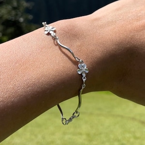 May include: A silver bracelet with a wavy design and small flower charms. The bracelet is on a person's wrist.