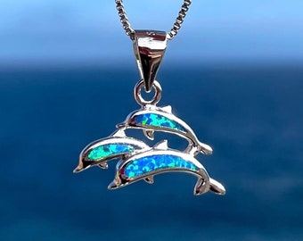 May include: A silver necklace with a pendant depicting three dolphins. The dolphins have blue and green opal-like inlays. The background shows a blurred view of the sea and sky.
