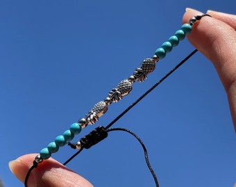 May include: A turquoise and silver beaded bracelet with pineapple charms. The bracelet is adjustable with a black cord and silver bead.